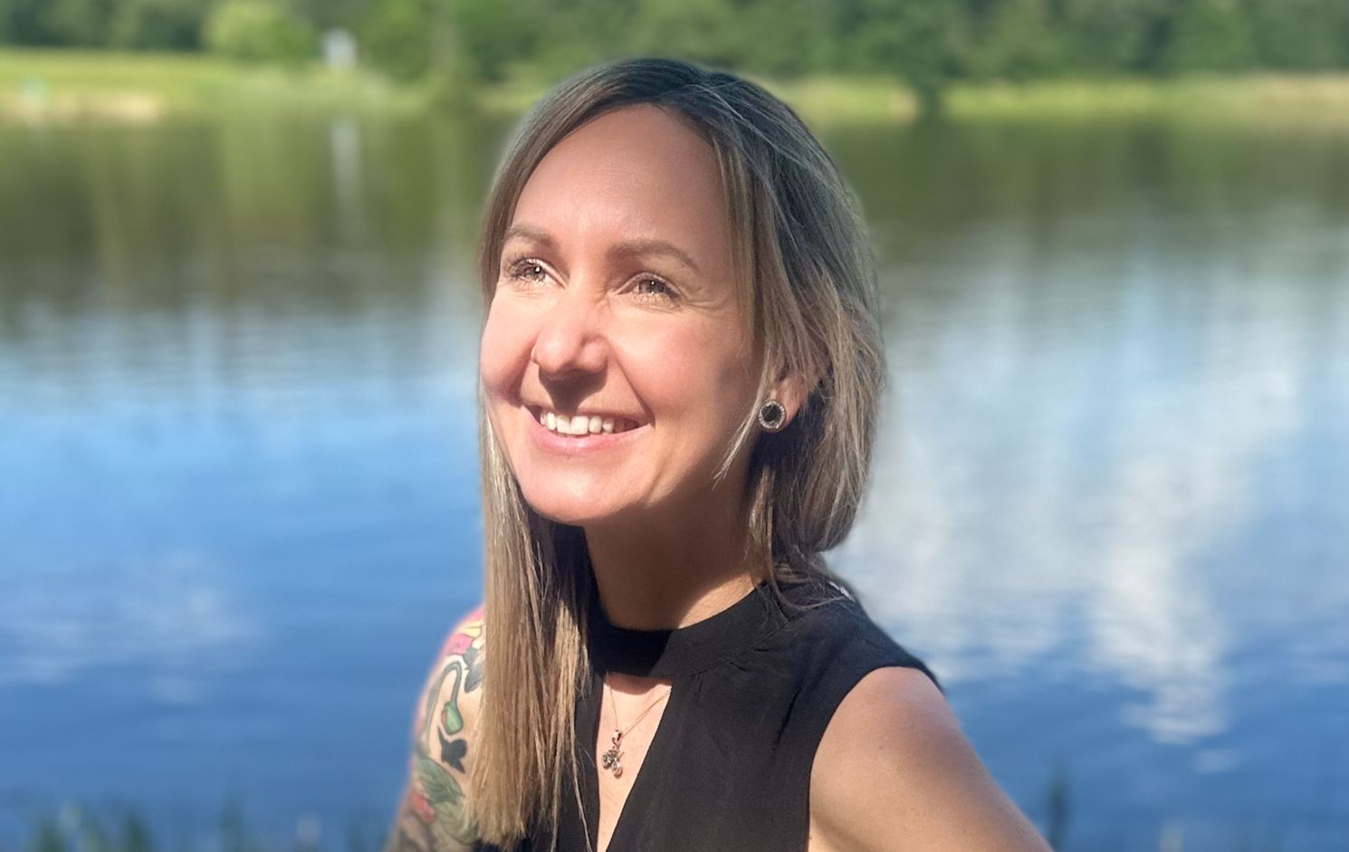 Woman smiling outdoors by a lake with trees in the background.